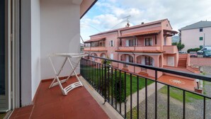 Un balcón con una silla plegable blanca, piso de tejas rojas y vista a un edificio rosa con varios balcones.