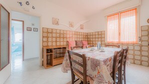 Comedor con una mesa dispuesta para cuatro personas, rodeada de sillas. Se observa un mueble de madera con estantes, una ventana con cortinas naranjas y una puerta que da a otra habitación.