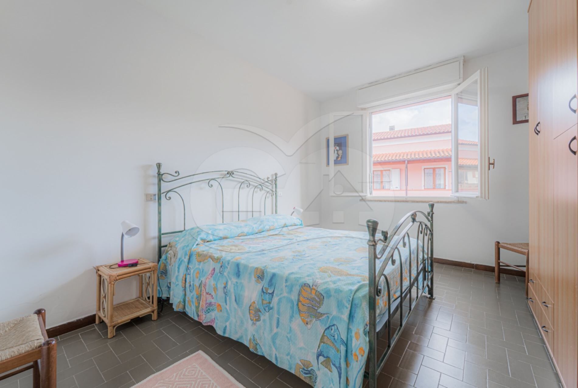 A bedroom with a bed, a small bedside table, a chair, a window with a view of buildings, and a wooden wardrobe.