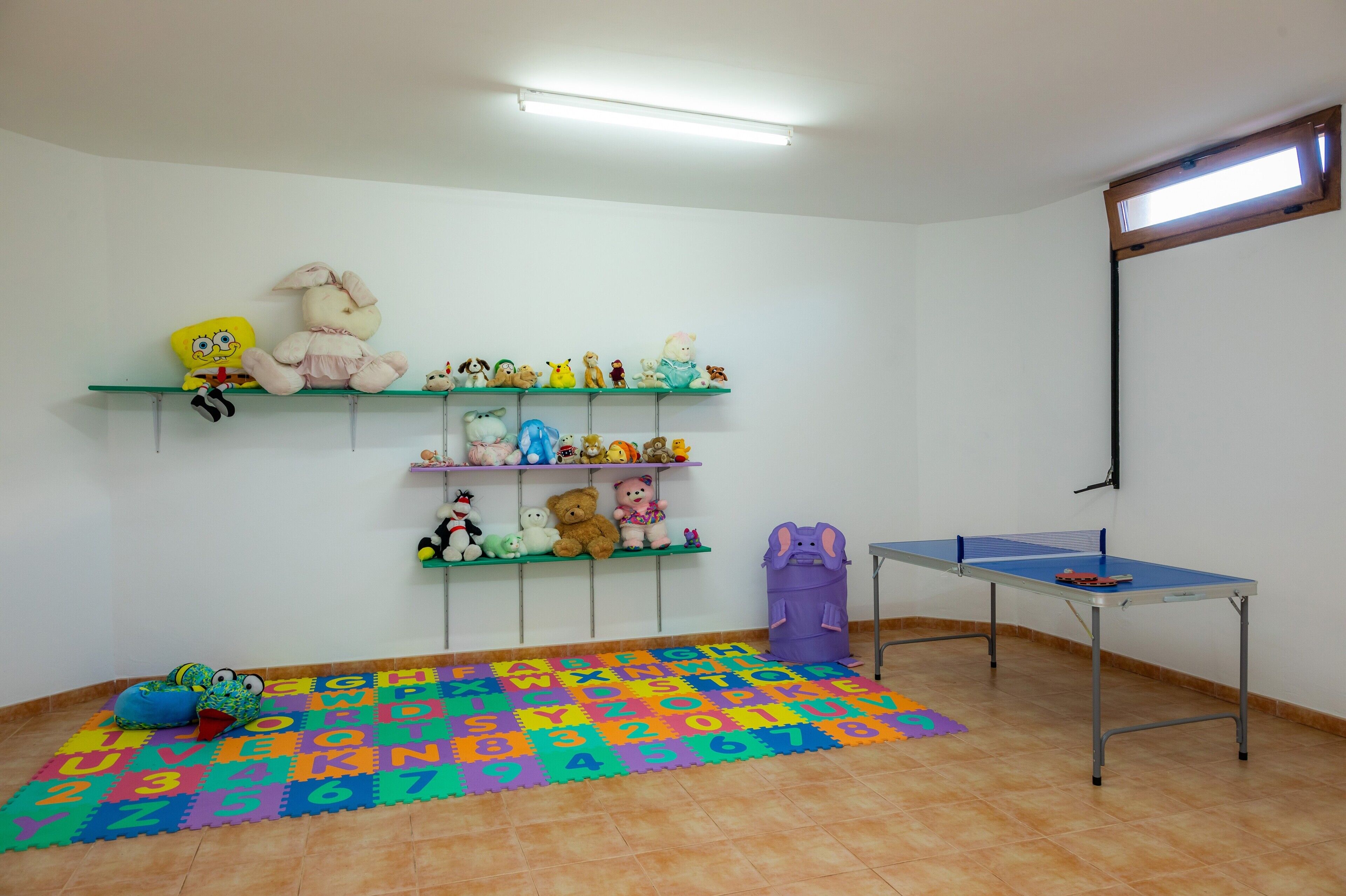 Een kamer met een tafeltennistafel, planken met knuffels, een kleurrijke lettermat en een raam.