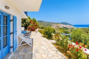 A stone-paved outdoor area with a table and chairs, overlooking a coastal town and the sea.