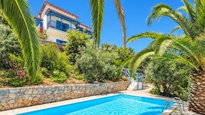 Une piscine bordée d’un mur en pierre et entourée de palmiers, le tout noyé dans une végétation luxuriante, avec un bâtiment aux accents bleus en arrière-plan.