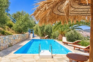 Una piscina con borde de piedra, sillas de descanso y una estructura de techo de paja.