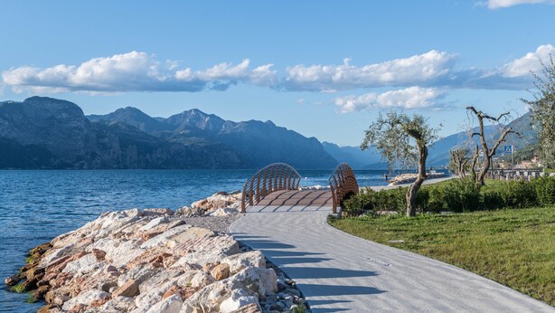 Ein gepflasterter Weg führt zu einer Brücke über ein Gewässer, im Hintergrund Berge.