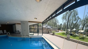 Ein überdachter Pool mit Glaswänden, einer Steinwand und Blick auf Sitzplätze im Freien und Bäume.