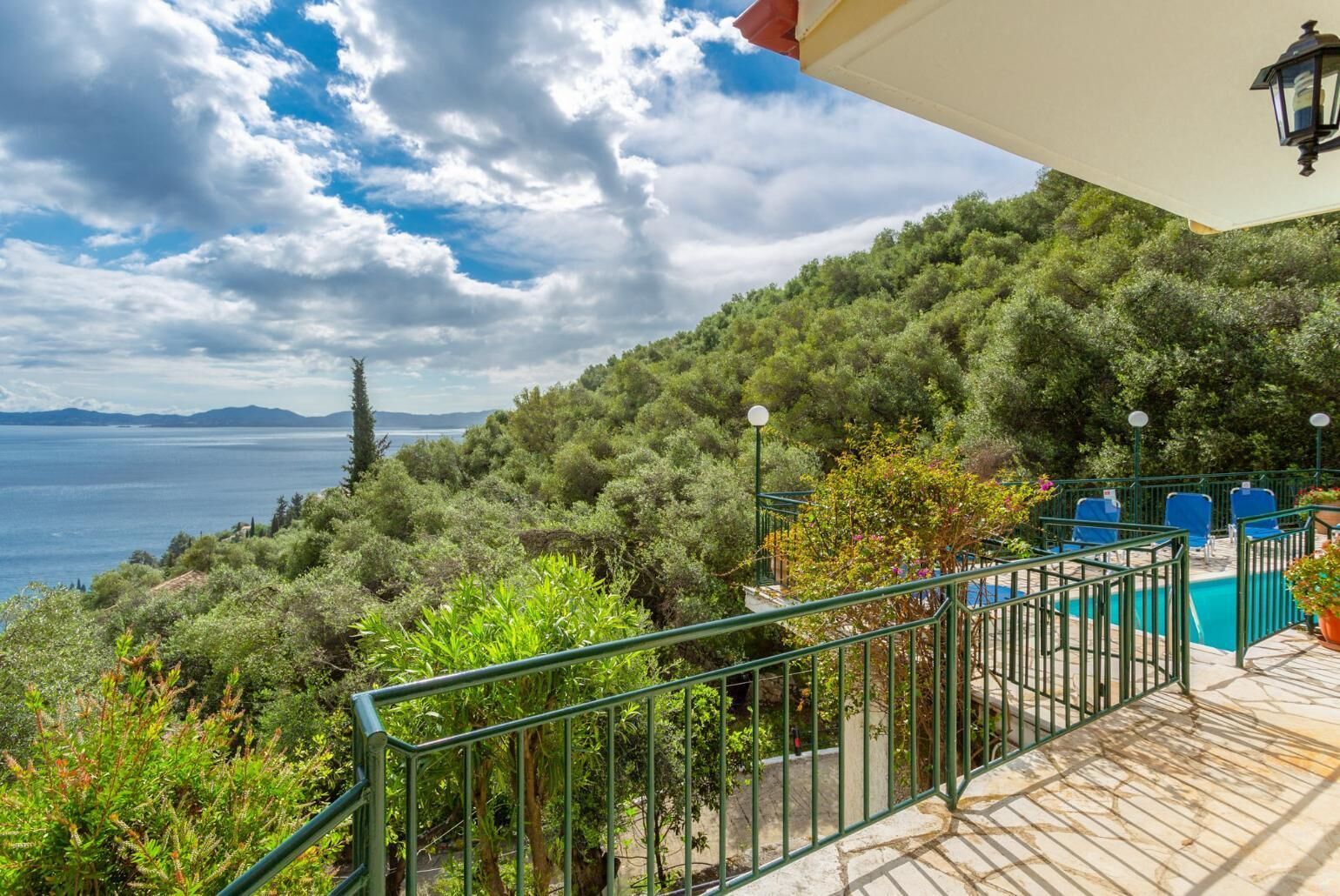 A terrace with a pool, surrounded by lush greenery and overlooking a body of water with mountains in the distance.