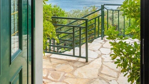 Un chemin de pierre mène à un balcon orné d’une rambarde en métal, d’où l’on peut admirer un plan d’eau et des montagnes lointaines sous un ciel partiellement nuageux.