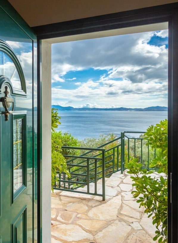 Un chemin de pierre mène à un balcon orné d’une rambarde en métal, d’où l’on peut admirer un plan d’eau et des montagnes lointaines sous un ciel partiellement nuageux.