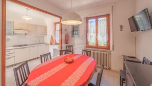 Un coin repas avec une table ronde recouverte d’une nappe rouge, des chaises, un saladier de fruits, une fenêtre avec des rideaux, un téléviseur accroché au mur et une cuisine en arrière-plan.