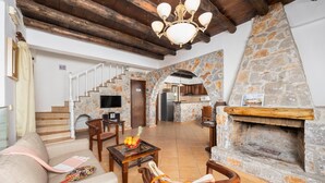 A spacious living area with a stone fireplace, wooden staircase, and a kitchen in the background. - Villa Tzina - Three Bedroom Villa, Sleeps 6 (Tavronitis)