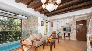 A spacious living room with a stone wall, a wooden staircase, a TV, and a pool visible through large windows. - Villa Tzina - Three Bedroom Villa, Sleeps 6 (Tavronitis)
