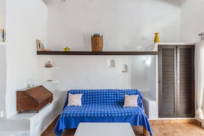 A room with a blue checkered sofa, a wooden chest, a shelf with books and a basket, and a window with shutters.