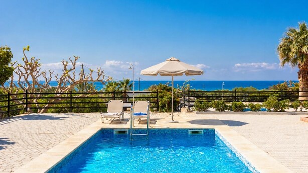 Piscina con dos sillas reclinables y una sombrilla, rodeada de una valla y palmeras, con vista al océano.