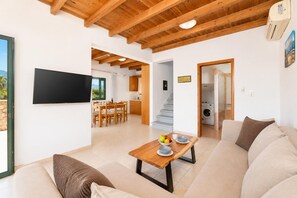 A modern living room with a sofa, a coffee table, and a TV. There is a dining area with a table and chairs. A kitchen is visible in the background.