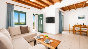 Una sala de estar con un sofá, una mesa de comedor de madera con sillas, un televisor montado en la pared y una ventana con cortinas.