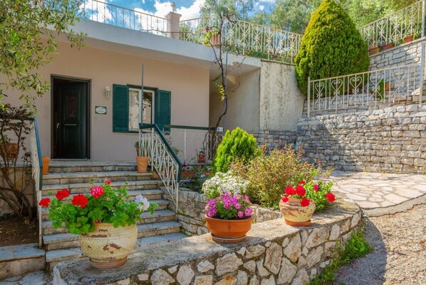 A stone staircase leading to a house with a balcony, surrounded by potted plants and flowers.