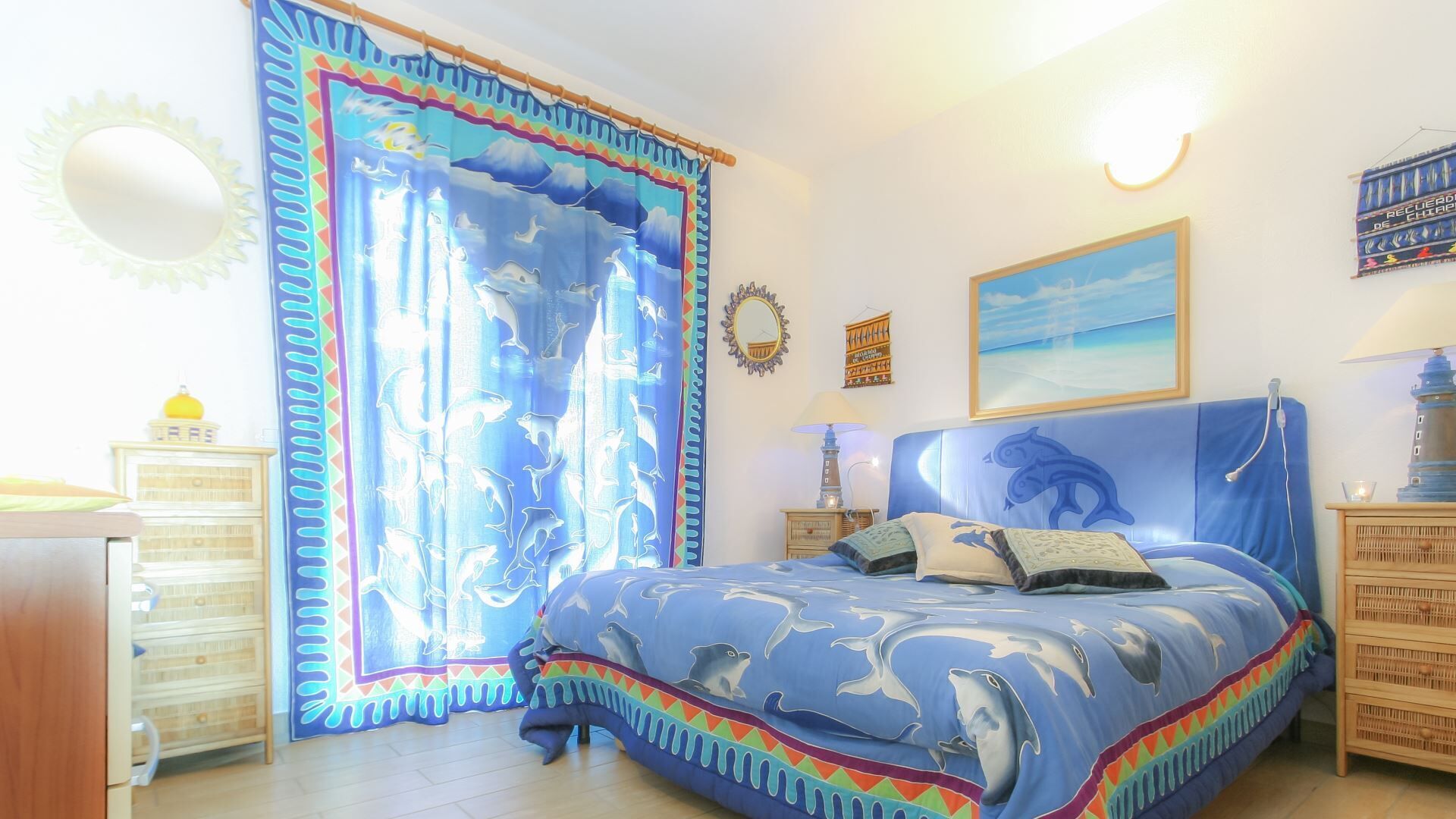 Une chambre à coucher avec un lit orné de motifs de dauphins, une grande fenêtre avec des rideaux, un miroir et une lampe fixée au mur.