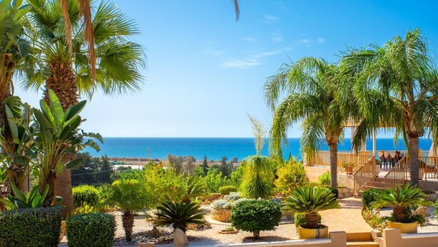 Un jardín ajardinado con palmeras y arbustos diversos, una pérgola de madera y vista al mar.