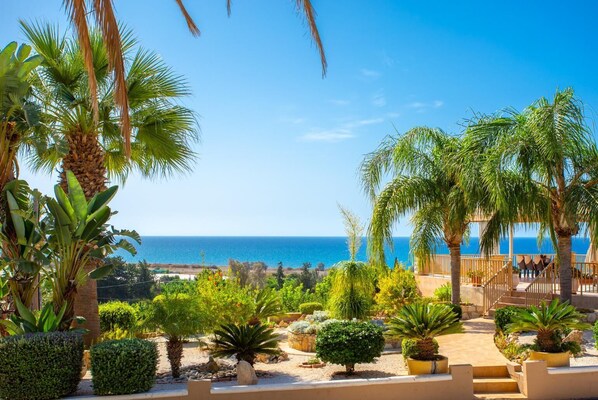 Un jardín ajardinado con palmeras y arbustos diversos, una pérgola de madera y vista al mar.