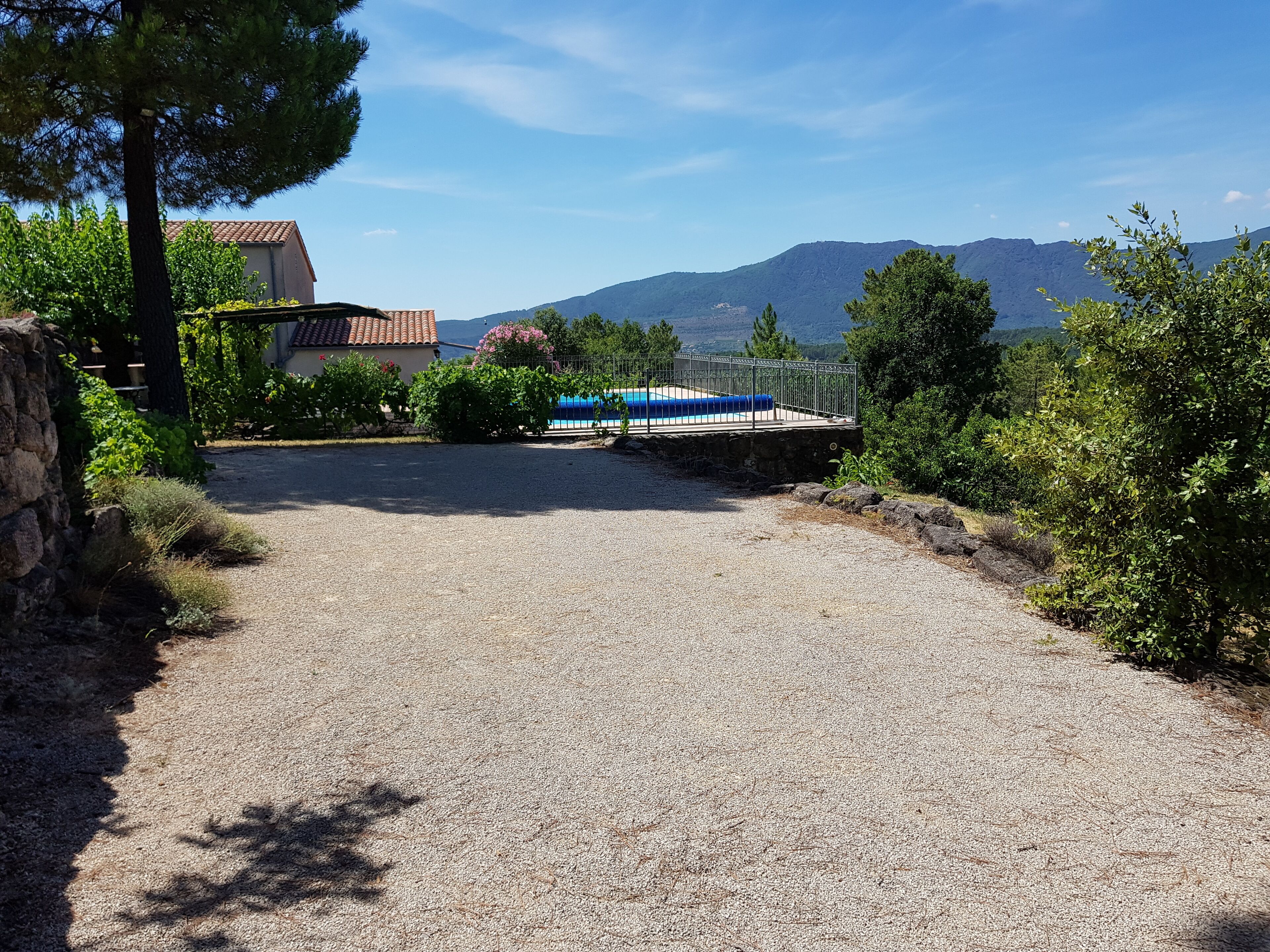 Un camino de grava que conduce a una casa con piscina, rodeada de follaje y montañas al fondo.