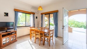 Een moderne woonkamer met een televisie, een houten eettafel en stoelen, grote ramen met gordijnen en een schuifdeur van glas naar een buitengebied.