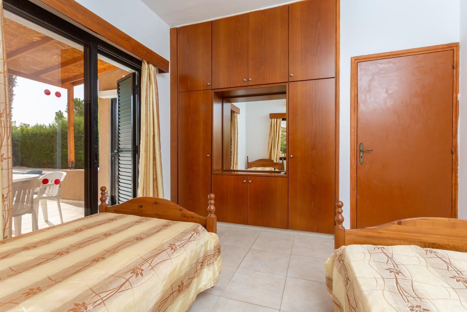A bedroom with two beds, wooden wardrobes, a large mirror, and a sliding glass door leading to an outdoor area.