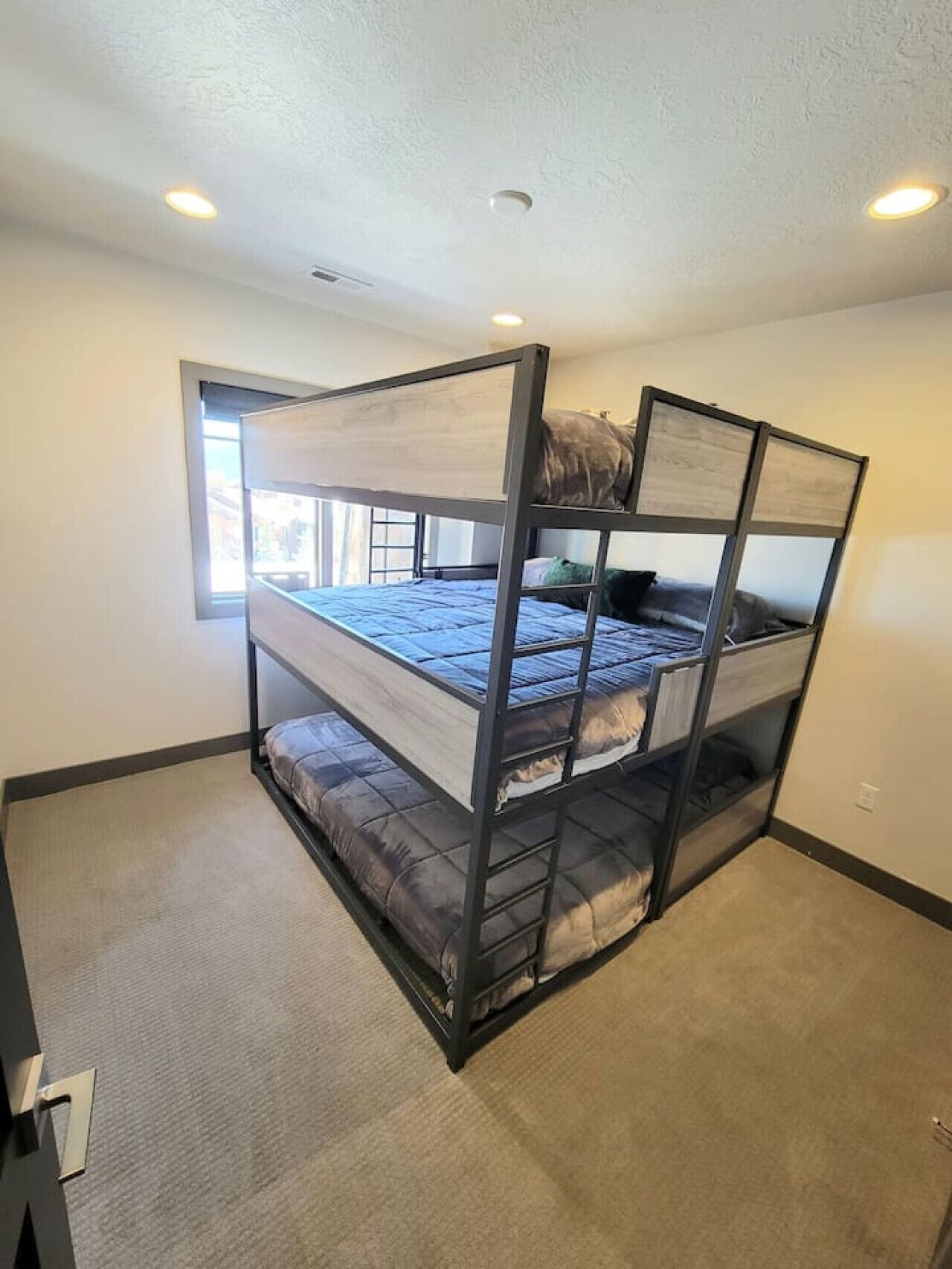 Cama litera con estructura de madera y colchón, ventana con persianas y una lámpara en el techo.