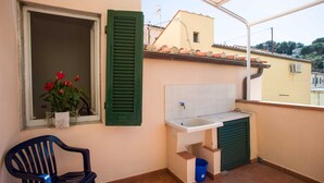 Un pequeño balcón con un lavabo, una silla y una ventana con contraventanas verdes.