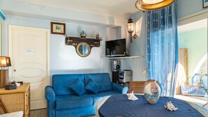 Una habitación con sofá azul, televisor, una mesita con lámpara y ventana con cortinas.