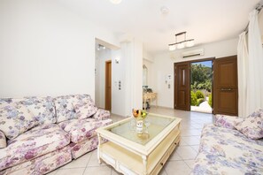 A living room with a floral sofa, a glass-top coffee table with a fruit bowl and wine glasses, and a view of a garden through an open door.