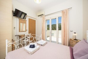 A bedroom with a bed, a television, a window with curtains, a bedside table with a lamp, and a bathroom with a mirror and towel rack.