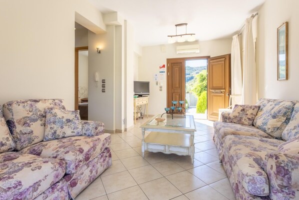 A living room with floral sofas, a glass coffee table, and a view of a kitchen and outdoor area.