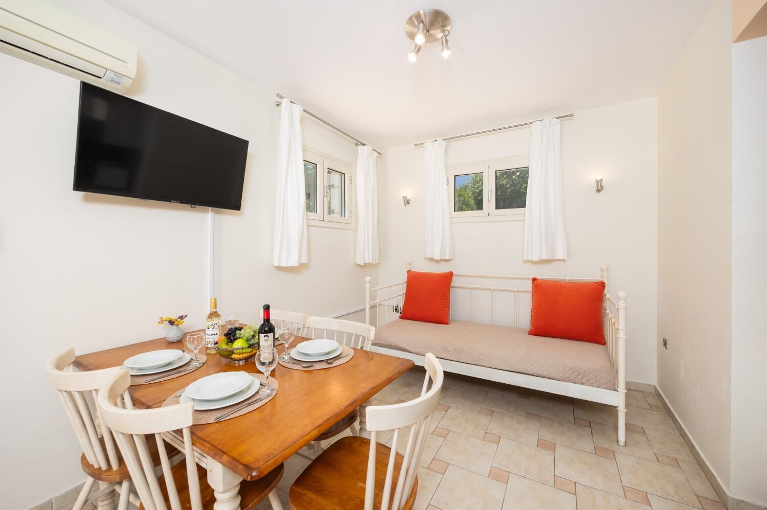 Un comedor con una mesa de madera dispuesta para dos personas, un televisor montado en la pared y un ventanal con cortinas.