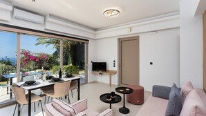 A modern living room with a dining area, large sliding glass doors, a flat-screen TV, and a sofa with cushions. - Villa Sevos - Two Bedroom Villa, Sleeps 4 (Crete)