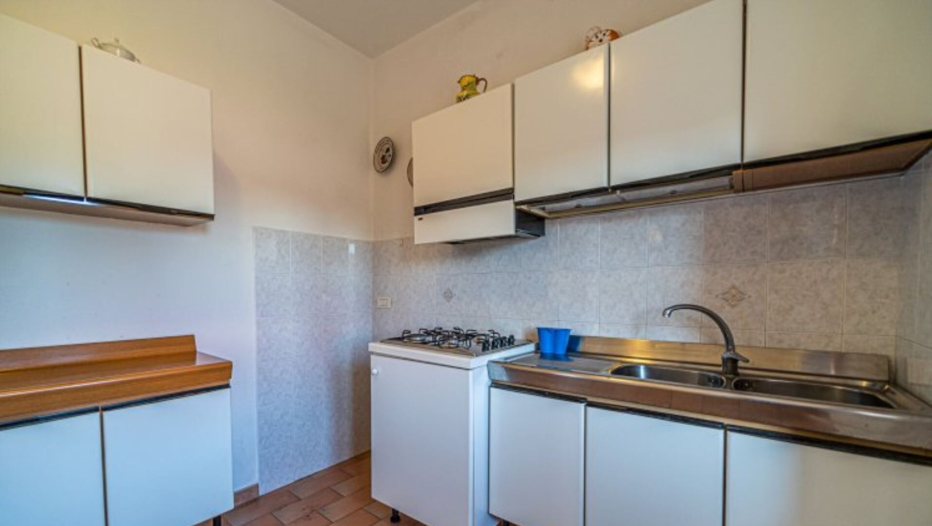 Une cuisine de taille réduite avec des meubles blancs, une cuisinière à gaz, un évier et une tasse bleue posée sur le comptoir.
