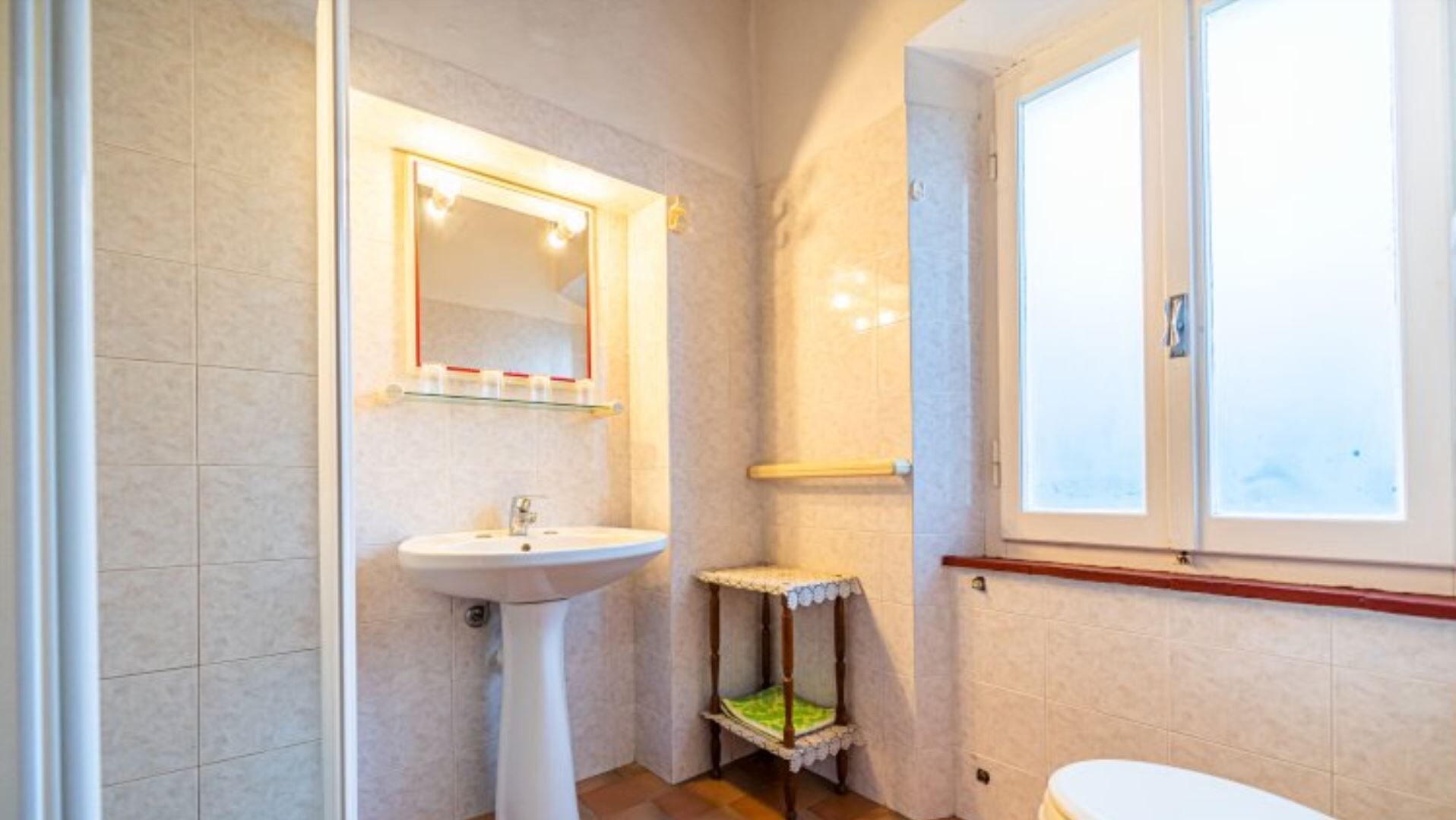 Une salle de bain avec un lavabo sur colonne, un miroir, une petite commode avec un coussin vert et des toilettes.