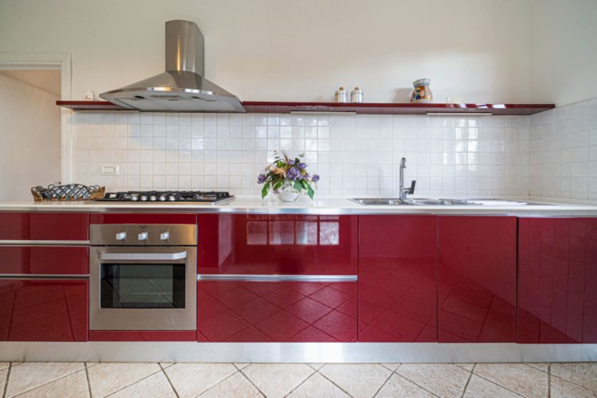 A kitchen with red cabinets, a stainless steel range hood, a built-in oven, and a white tiled backsplash.