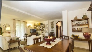 A cozy living room with a wooden dining table, a sofa, and a TV. There are decorative items on the walls and a bookshelf filled with books and knick-knacks. - Villa Monserrato with garden and sea view (Porto Azzurro)