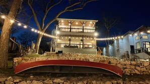 A two-story building with a stone bridge in front, surrounded by trees and string lights. - River Repose Rr 7134 4 Bedroom Home by RedAwning (New Braunfels)