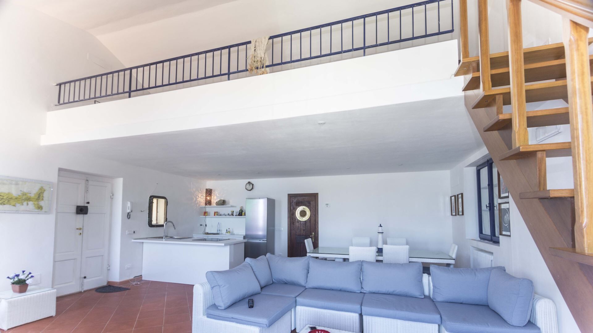 Amplia sala de estar con un sofá seccional grande, una cocina con refrigerador, una mesa de comedor y una escalera que lleva a un nivel superior.