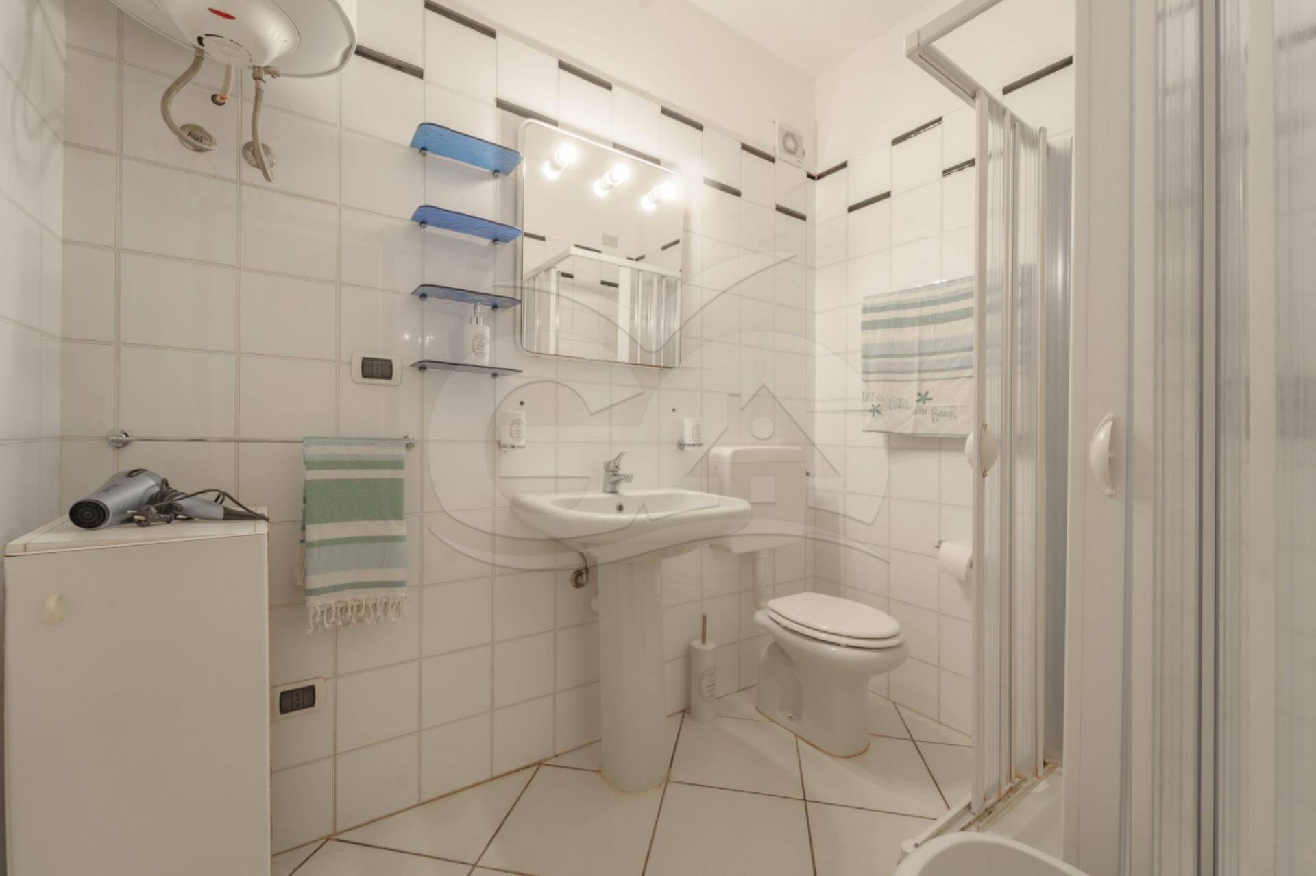 Une salle de bain comprenant un lavabo, des toilettes et un espace douche. On y trouve un porte-serviettes et un sèche-cheveux, qui sont posés sur un meuble.