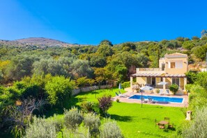 Une maison à deux étages avec une piscine, entourée d’une végétation luxuriante et de collines.