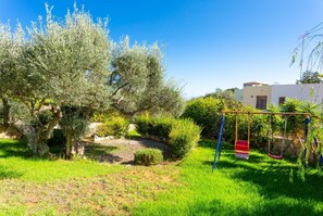 A garden with olive trees, a swing set, and a white building in the background.