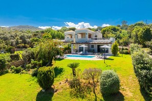 A two-story house with a swimming pool, surrounded by lush greenery and hills in the background.