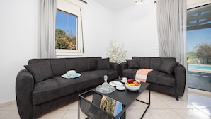 A modern living room with two dark sofas, a coffee table with fruit and a bowl, a vase with flowers, and a view of a pool through a large window. - Villa Georgia - Three Bedroom Villa, Sleeps 6 (Crete)