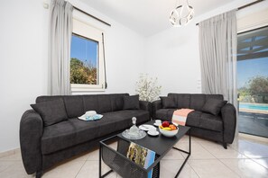 Ein modernes Wohnzimmer mit zwei dunklen Sofas, einem Couchtisch mit Obst und einer Schale, einer Vase mit Blumen und Blick auf einen Pool durch ein großes Fenster.