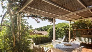 Un patio techado al aire libre con mesa y sillas, rodeado de vegetación y vista a una casa en el fondo.