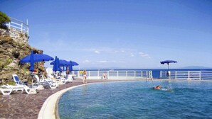 Zona junto a la piscina con sillas reclinables y sombrillas, vista despejada del mar y un acantilado rocoso.