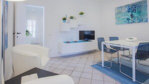Une pièce moderne et bien éclairée, avec un canapé blanc, une table à manger et des chaises, un téléviseur posé sur un meuble blanc et un tableau accroché au mur.