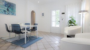 Une pièce moderne et bien éclairée, avec un coin repas comprenant une table blanche et des chaises bleues, un canapé blanc et une grande fenêtre avec des rideaux.